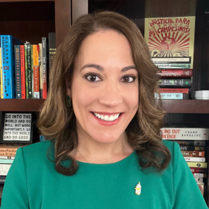 一名棕色长发及肩、身穿绿色衬衫的女子微笑着站在摆满书籍的书架前。