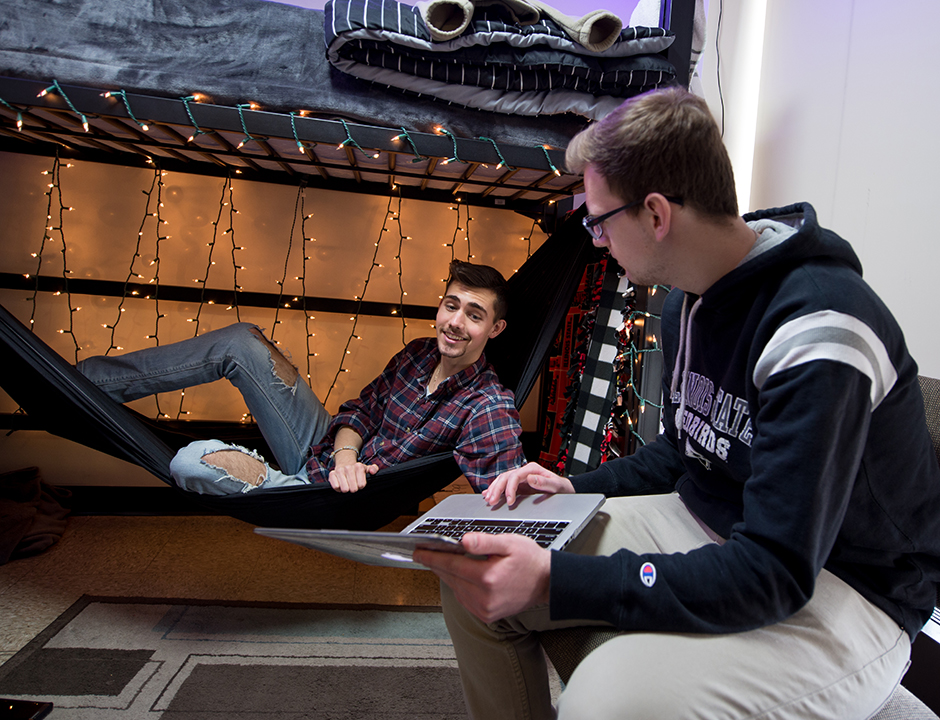 两个男孩在一个学生宿舍,一分之一吊床阁楼下床,另一个坐在桌子椅子和一台笔记本电脑。