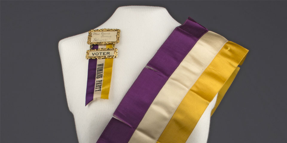 白色礼服，身穿紫、白、黄三色全国妇女党绶带，丝带上写着“戴维斯尤因夫人伊利诺伊州选民妇女党”。