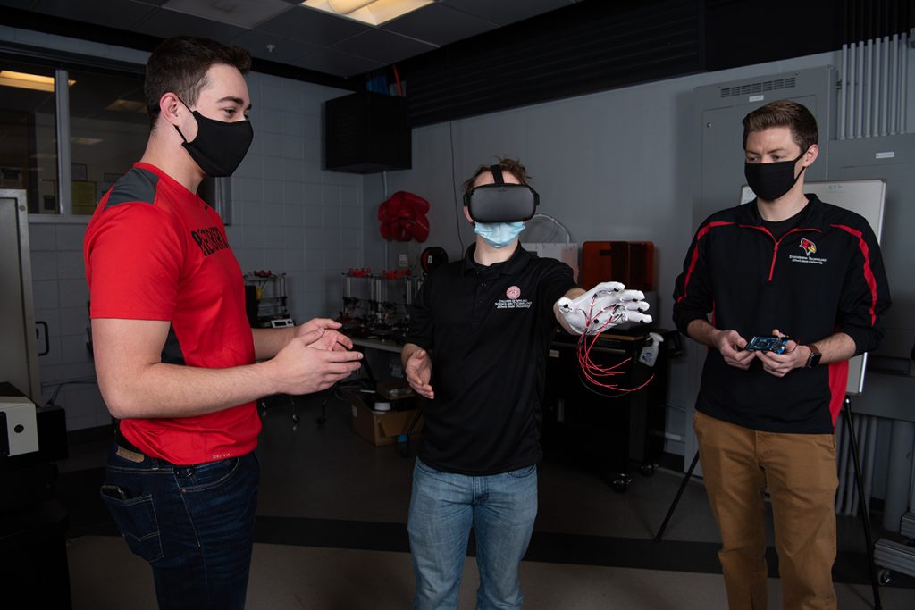 Jake Weihe站在最左边。Alex Diffor站在中间，戴着VR头盔，一只手戴着触觉手套，伸出在他面前。乔丹·奥斯本站在右边，手里拿着一块小电路板。