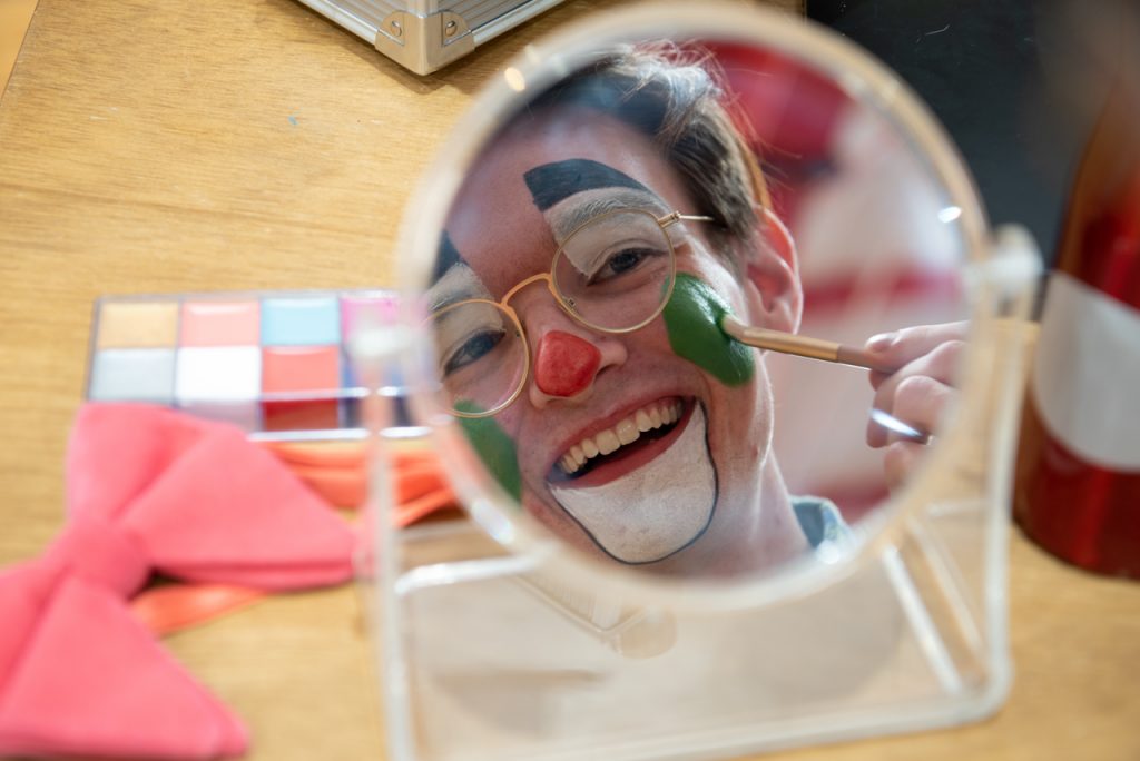 乔恩·富尔顿在镜子里的倒影。富尔顿化着小丑的妆，用化妆刷在脸颊上画了一个绿色的圆圈。