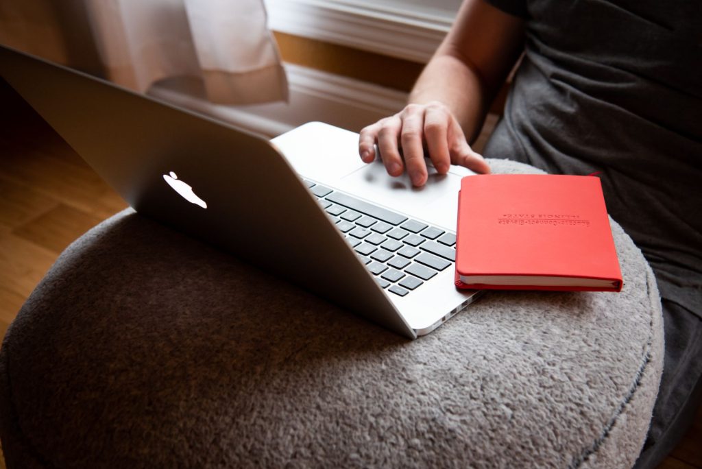 一台打开的苹果MacBook放在软垫桌子上，一个红色笔记本电脑平衡在键盘边缘，一个人用手指使用触控板