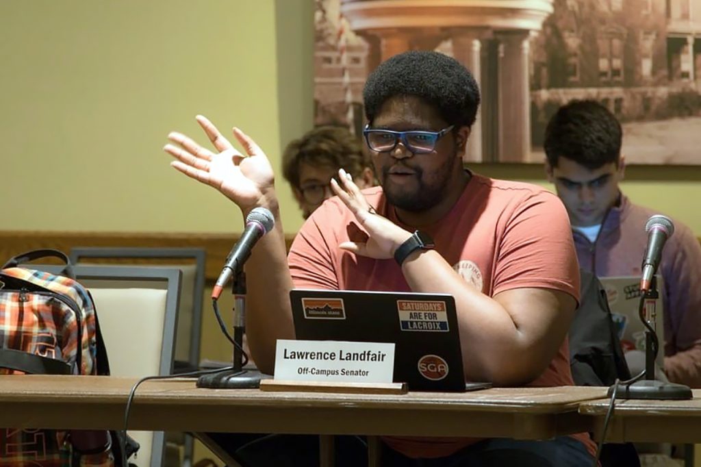 劳伦斯Landfair对麦克风讲话,坐在一张桌子前,身后的一台笔记本电脑