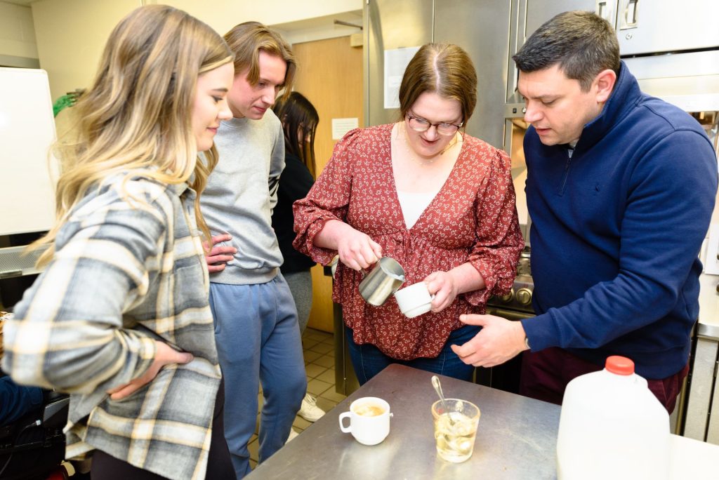 两名学生和Erol Sozen博士正在观察另一名学生将起泡牛奶倒入拿铁咖啡中。