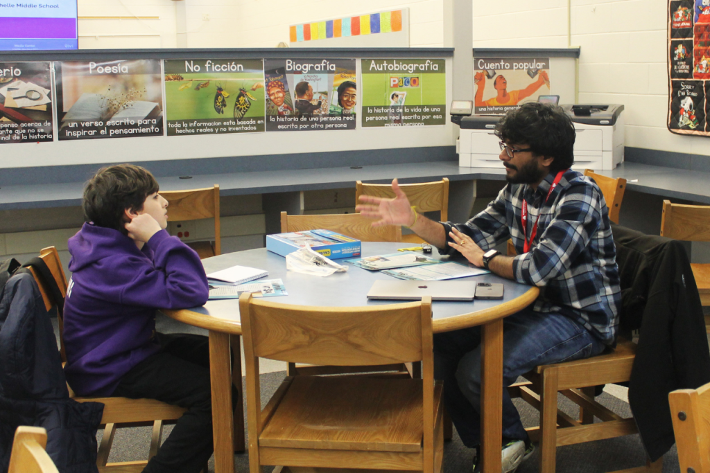 一个男孩和一个年轻人坐在图书馆的一张桌子旁，他们之间摊开了色彩鲜艳的作业纸。这名男子在向男孩解释一个文学概念时，用手比划着。