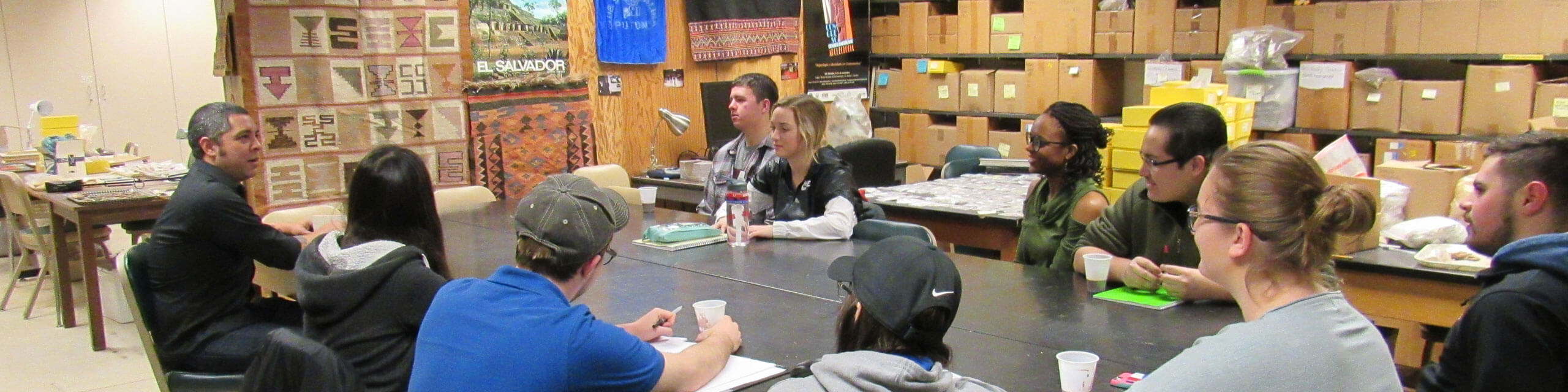 一个人类学小班学生围坐在一张桌子旁，聚精会神地听着教授讲课。
