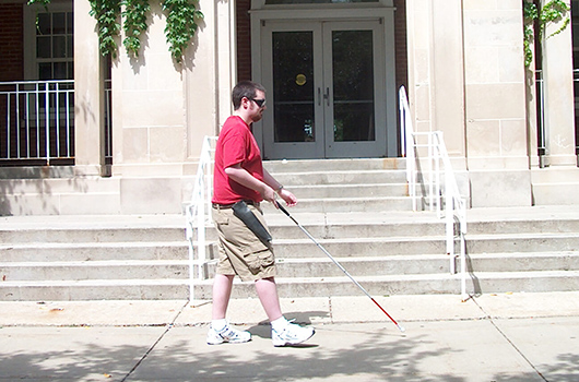一个拄着白色拐杖的学生走在费尔大厅外面。