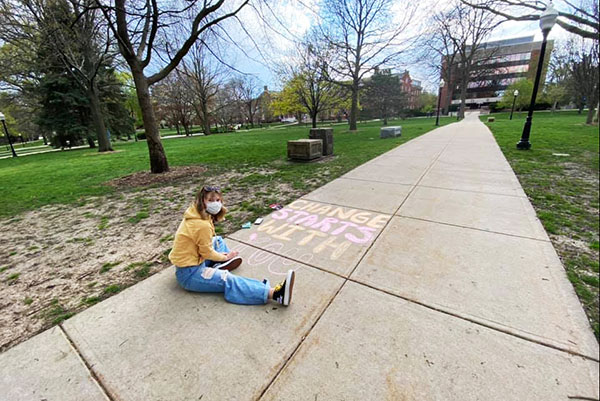一名学生坐在人行道上，用粉笔写下“改变从你开始”。